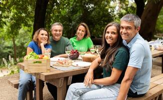 A group of five friends gathers around a picnic table in a verdant park, sharing food and laughter on a warm afternoon, with fruits and snacks spread out before them. photo