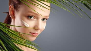 Red-Haired Girl With Facial Moisturizer Cream Posing Looking Through Green Leaves In Studio Over White Background. Face Beauty Products, Natural Skincare Cosmetics Concept photo