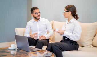 A man and woman engage in a business discussion while seated on a couch, enjoying coffee in a modern lounge setting, focused on exchanging ideas and insights. photo