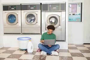A young man sits on the floor of a laundromat, focused on his laptop, with laundry machines in the background. He appears relaxed and productive as he multitasks while waiting for clothes. photo