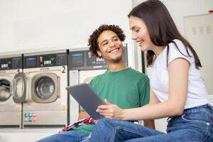 Two young adults sit side by side in a laundromat, smiling and engaging with a tablet as they wait for their laundry to finish in the background. photo