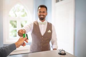 A man with a bow tie is handing a credit card to a person at a hotel reception desk. A bell is in the foreground on the counter. photo