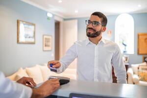 A man greets the receptionist at the hotel desk, handing over a key card while smiling. The bright, inviting lobby features comfortable seating and warm decor, creating a welcoming atmosphere. photo