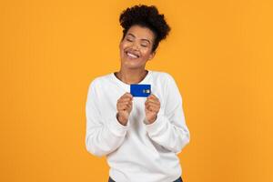 A smiling young woman wearing a white sweater confidently displays a blue card in front of a vibrant orange backdrop, radiating happiness and enthusiasm. photo