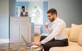 A man in a white shirt sits on a comfortable couch, focused on his laptop, sipping coffee. A second person is in the background by the reception area, creating a professional ambiance. photo