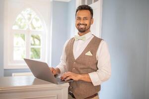 A man with a beard, wearing a white shirt, brown vest, and light green bow tie, receptionist smiles while looking at the camera. He is standing in front of a window with a white frame at hotel desk photo