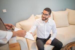 A man in a white shirt with a beard and glasses is sitting on a white couch. He is looking to the side, smiling. An unseen person is handing him a document, which he is accepting with his left hand. photo