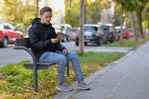A man chatting texting message using a mobile smartphone device sitting on a bench at the street on a sunny autumn day photo