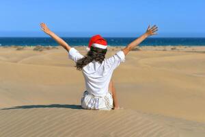 A girl in a dune wearing a Santa hat with hands up. Back view. The concept of a Christmas vacation in hot countries, at the sea. Dune near the sea in Maspalomas, Gran Canaria photo