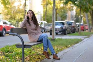A woman talking on a phone sitting on a bench at the street on a sunny autumn day. A woman smiles while using a mobile smartphone device photo