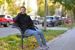 A man talking on a phone sitting on a bench at the street on a sunny autumn day. A man uses a mobile smartphone device photo