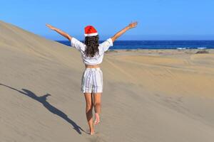 A girl in a dune wearing a Santa hat with hands up. Back view. The concept of a Christmas vacation in hot countries, at the sea. Dune near the sea in Maspalomas, Gran Canaria photo