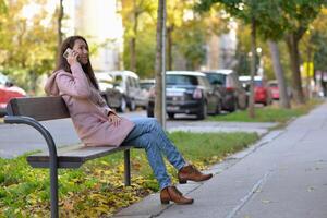 A woman talking on a phone sitting on a bench at the street on a sunny autumn day. A woman smiles while using a mobile smartphone device photo