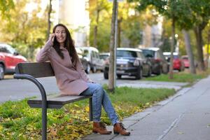 A woman talking on a phone sitting on a bench at the street on a sunny autumn day. A woman smiles while using a mobile smartphone device photo
