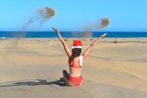 A girl in a dune wearing a Santa hat and a red swimsuit throws sand up. Back view. The concept of a Christmas vacation in hot countries, at the sea. Dune near the sea in Maspalomas, Gran Canaria photo