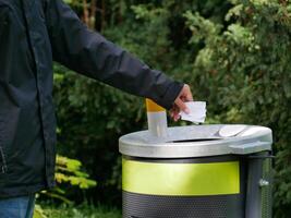 A man throws trash into a rubbish can in the park. Close up hand throwing paper in bin. A concept of ECO friendly Earth care and protection photo