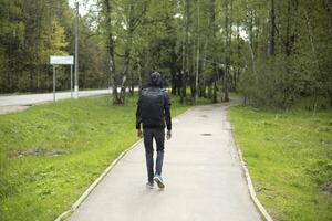 Guy walks through park in summer. Man in black clothes with backpack on his back. Student goes to school on street. photo