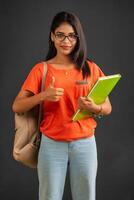 Beautiful young girl with a backpack standing and holding a notebook, posing on a grey background. photo