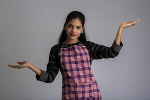 Portrait of Indian woman chef or cook in an apron, presenting, pointing, with ok sign, thumbs up on the grey background. photo