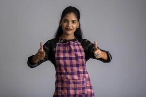 Portrait of Indian woman chef or cook in an apron, presenting, pointing, with ok sign, thumbs up on the grey background. photo