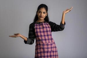 Portrait of Indian woman chef or cook in an apron, presenting, pointing, with ok sign, thumbs up on the grey background. photo