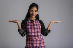 Portrait of Indian woman chef or cook in an apron, presenting, pointing, with ok sign, thumbs up on the grey background. photo