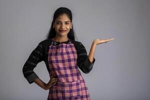Portrait of Indian woman chef or cook in an apron, presenting, pointing, with ok sign, thumbs up on the grey background. photo