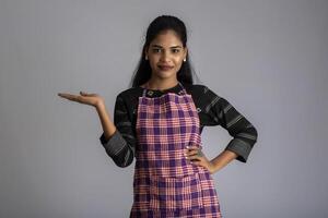 Portrait of Indian woman chef or cook in an apron, presenting, pointing, with ok sign, thumbs up on the grey background. photo