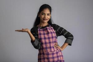 Portrait of Indian woman chef or cook in an apron, presenting, pointing, with ok sign, thumbs up on the grey background. photo