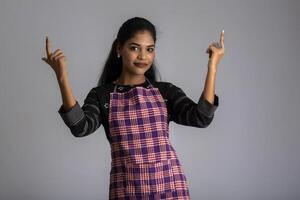 Portrait of Indian woman chef or cook in an apron, presenting, pointing, with ok sign, thumbs up on the grey background. photo