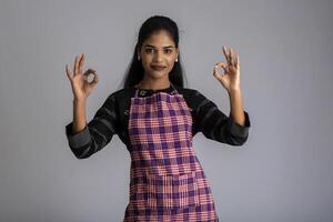 Portrait of Indian woman chef or cook in an apron, presenting, pointing, with ok sign, thumbs up on the grey background. photo