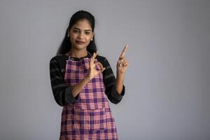 Portrait of Indian woman chef or cook in an apron, presenting, pointing, with ok sign, thumbs up on the grey background. photo