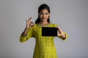 Young beautiful girl holding and showing blank screen of smartphone or mobile or tablet phone on a gray background. photo