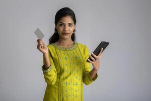 Young smiling beautiful woman or girl presenting credit card while using mobile or smartphone on grey background. photo