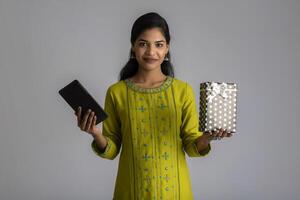 Portrait of young happy smiling Girl holding gift box and showing smartphone or tablet on a grey background. photo