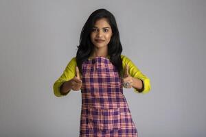 Portrait of Indian woman chef or cook in an apron, presenting, pointing, with ok sign, thumbs up on the grey background. photo