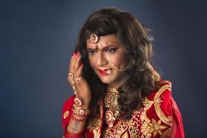 Young crossdresser man wearing Indian wedding clothes of a bride with bridal makeup and posing on grey background, looking fashion and glamorous. photo