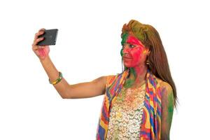 Young girl with colourful face taking selfie using smartphone on Holi festival. Festival and technology concept on white background. photo