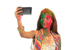 Young girl with colourful face taking selfie using smartphone on Holi festival. Festival and technology concept on white background. photo