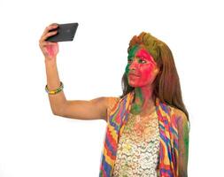 Young girl with colourful face taking selfie using smartphone on Holi festival. Festival and technology concept on white background. photo