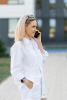 Young woman in a white linen suit, talking on a cell phone, walking on a city street in the summertime photo