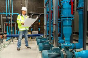 asian man worker in protective uniform and with hardhat using tablet for checking temperature in pipes Factory interior. photo
