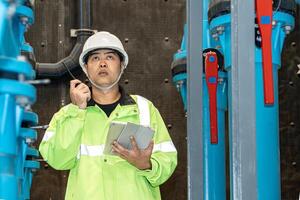 asian man worker in protective uniform and with hardhat using tablet for checking temperature in pipes Factory interior. photo