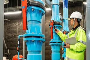 asian man worker in protective uniform and with hardhat using tablet for checking temperature in pipes Factory interior. photo
