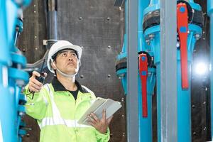 asian man worker in protective uniform and with hardhat using tablet for checking temperature in pipes Factory interior. photo
