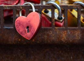heart with a keyhole is hanging from a rusty metal fence. The heart is red and has a shiny, metallic appearance. The chain is also metallic photo