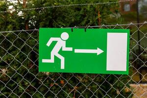 A green exit sign with a white stick figure running right is attached to a chain-link fence photo