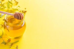A jar of liquid honey from Linden flowers and a stick with honey on yellow background. Copy space photo