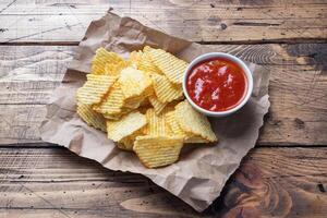 Spiced potato chips on paper and a plate of ketchup sauce on a wooden table. copy space. photo
