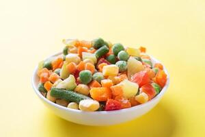 Chopped frozen vegetables in a plate on a yellow background. Corn peas pepper carrots. copy space. photo